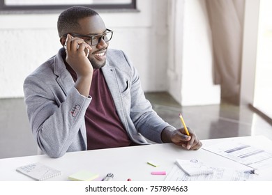 Cheerful black plump male enterpreneur has happy expression, discuss conference with colleague via smart phone, satisfied with company`s review in press, rejoice successful investment of money - Powered by Shutterstock