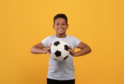 Cheerful Black School Boy Holding Soccer Ball And Smiling At Camera, Playing Football, Yellow Studio Background, Copy Space. Sport, Leisure, Entertainment, Hobby For Kids Concept