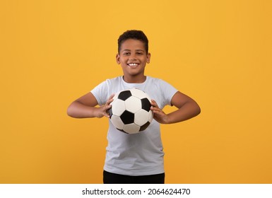 Cheerful black school boy holding soccer ball and smiling at camera, playing football, yellow studio background, copy space. Sport, leisure, entertainment, hobby for kids concept