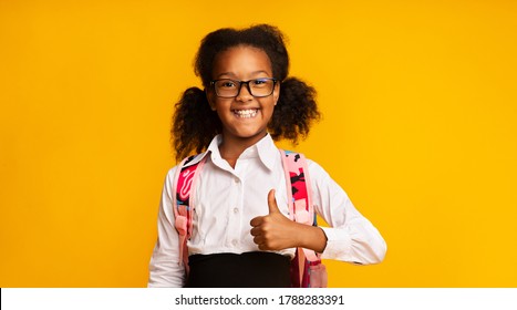 Cheerful Black School Girl Gesturing Thumbs-Up Smiling To Camera Posing Over Yellow Studio Background. I Like School Concept. Panorama