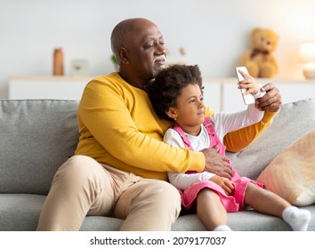 Cheerful Black Small Girl And Old Man Hugging And Taking Selfie On Smartphone On Sofa In Living Room Interior. Selfie Of Grandfather And Granddaughter, Video Call During Covid-19 Lockdown, New App