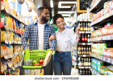 Cheerful Black Spouses Choosing Food In Grocery Store Walking With Cart Full Of Products Along Supermarket Aisles. African American Family Buying Groceries Together In Shop Indoor - Powered by Shutterstock