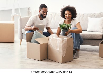 Cheerful Black Spouses Packing Moving Boxes Putting Plants In Cardboard Box Preparing For Relocation Together Sitting Indoors. New Home, Real Estate Purchase And Family Housing Concept
