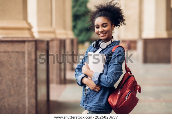Cheerful Black Student Girl Hugging Books Posing\
With Backpack Near College Building Outdoor, Smiling To Camera.\
Modern Education, College Tuition And Grants, Studentship And Study\
Abroad Concept