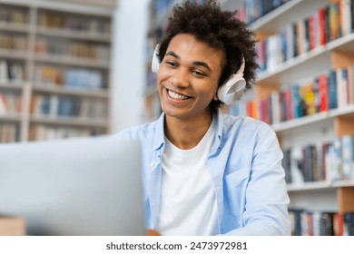 Cheerful Black student man in wireless headphones looking at laptop and smiling, talking on video call from college library