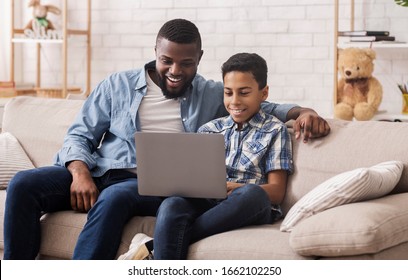 Cheerful Black Teen Boy And His Dad Watching Comedy Movie On Laptop Together, Relaxing On Sofa In Livivng Room, Empty Space