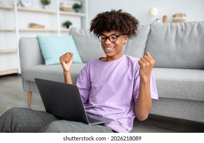 Cheerful Black Teen Guy Sitting On Floor With Laptop, Making YES Gesture, Getting Good News On Web, Achieving Success, Celebrating Victory At Home. Online Life During Covid Lockdown Concept