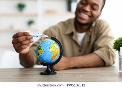 Cheerful Black Traveler Guy Playing With Airplane Toy Model And World Globe Sitting At Desk At Home. Vacation Planning, Touristic Destinations Concept. Selective Focus, Shallow Depth