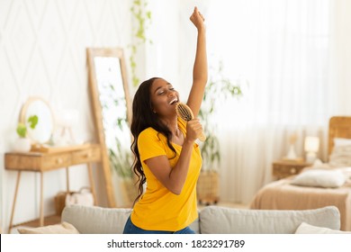 Cheerful black woman with hairbrush as mic singing her favorite song and dancing at home. Millennial African American lady pretending to be famous singer, having fun in living room - Shutterstock ID 1823291789