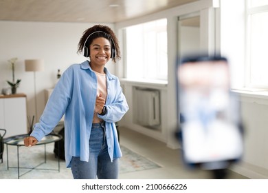 Cheerful black woman in headphones dancing at camera filming video using phone on tripod at home, creating trendy content on mobile app to share on social media, selective focus on millennial lady