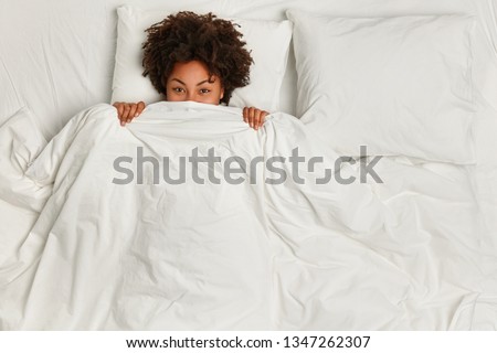 Cheerful black woman hides under soft white blanket, has fun in bed, rejoices good morning, has curly hair, rests in bed. View from above. Copy space. People, ethnicity and sleeping concept.