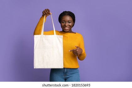 Cheerful Black Woman Holding And Pointing At Blank Textile Tote Eco Canvas Shopping Bag While Posing Over Purple Studio Background, Looking At Camera, Showing With Finger At Copy Space For Your Logo