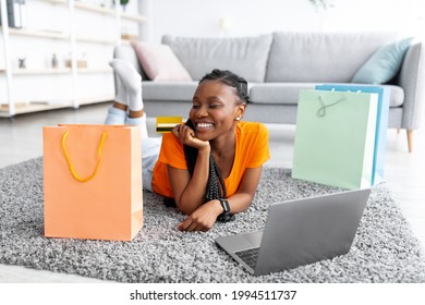 Cheerful Black Woman With Laptop Pc And Credit Card Lying On Floor, Surrounded By Shopping Bags At Home. Happy African American Lady Purchasing Over Internet At Living Room During Coronavirus