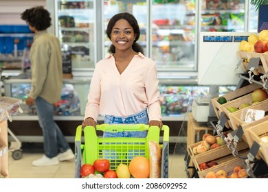 Cheerful Black Woman Shopping Grocery Products Posing With Shop Cart Smiling To Camera Standing In Supermarket. Shot Of Happy Customer Buying Food In Groceries Store