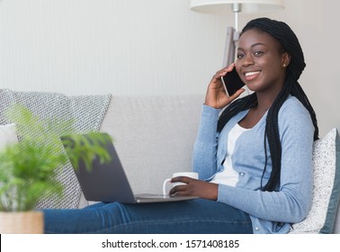 Cheerful Black Woman Talking On Phone, Drinking Morning Coffee And Using Laptop At Home