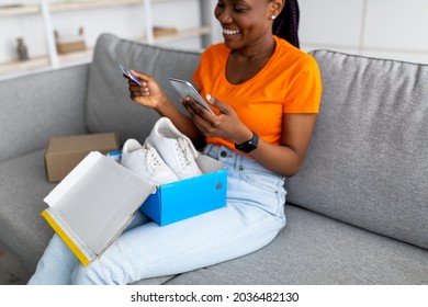 Cheerful black woman unpacking parcel with new sports shoes, holding credit card and smartphone, writing online store feedback, indoors. Happy Afro lady shopping on web, satisfied with good service Cheerful black woman unpacking parcel with new sports shoes, holding credit card and smartphone, writing online store feedback, indoors. Happy Afro lady shopping on web, satisfied with good service