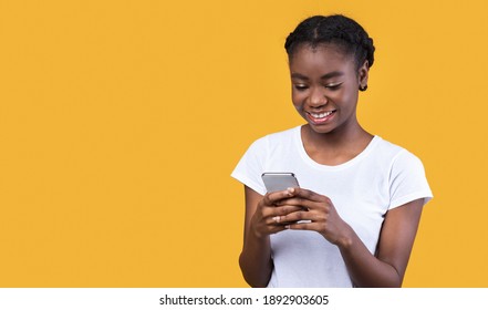 Cheerful Black Woman Using Phone Texting And Networking Standing Over Yellow Studio Background. African Young Lady Using Mobile Application On Smartphone. Empty Space For Text