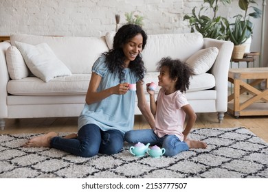 Cheerful Black Young Mother And Little Daughter Kid Having Fun, Playing Tea Party, Pretending Drinking Coffee From Toy Cups, Playing Funny Role Games On Warm Heating Floor In Cozy Living Room