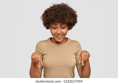 Cheerful black young woman points down, notices something amazing on floor, has positive smile, crisp hair, wears casual beige clothes, advertises her new shoes, models against white studio wall