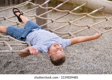 Cheerful Blond Boy Smiling And Lying On A Rope Swing In Public Park.