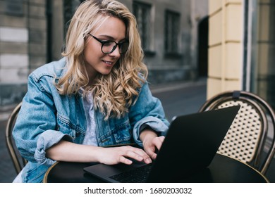 Cheerful blond lady in denim jacket and spectacles smiling and typing on laptop keyboard while sitting at restaurant table on blurred background of city street
