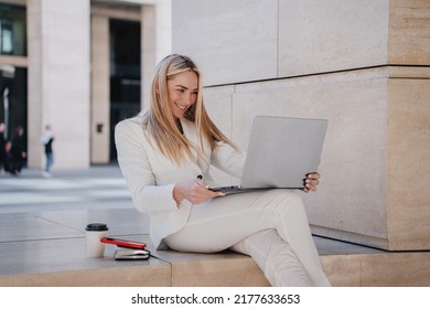 Cheerful blonde businesswoman sitting outdoors with laptop , looking at screen happily . Smiley student in white suit making video call. Education and finance. Gorgeous  swedish woman working outside.