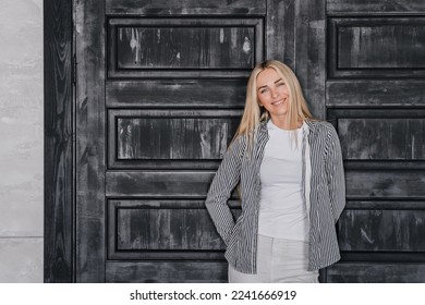 Cheerful blonde Caucasian young woman in casual toothy smiles stands against door at home. Pretty model with folded hands in grateful expression looks at camera happily. Successful people. - Shutterstock ID 2241666919