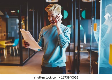 Cheerful Blonde Chinese Male Entrepreneur Reading Document And Having Mobile Phone Conversation In Office, Skilled Asian Businessman Making Banking Via Smartphone Call Holding Papers With Transactions