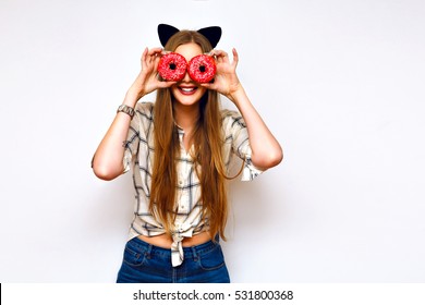 Cheerful blonde fashion girl with black sexy cat ears holding bright pink tasty donuts against her eyes. Cute smile. Delicious colorful dessert. Diet. Fast food. Isolated.