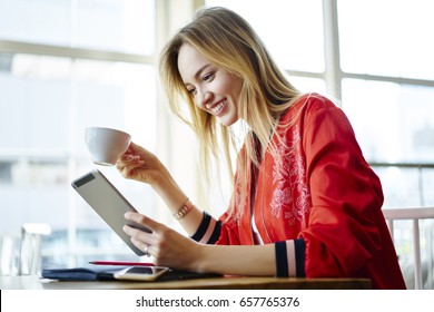 Cheerful Blonde Student Drinking Coffee While Reading Electronic Book On Tablet Connected To Wifi In Cafe,young Hipster Girl Sharing Multimedia Files And Blogging In Social Networks In Good Mood  