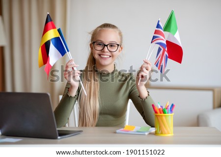 Cheerful blonde teen girl with glasses sitting at workdesk with international flags, home interior, using laptop, studying foreign languages. International education, online language courses