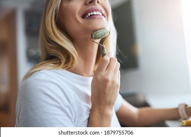 Cheerful Blonde Woman Doing Anti Age And Lifting Massage On Chin And Neck With Modern Roller Massager With Jade Stone In Room Indoors