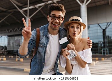 Cheerful blonde woman in hat makes funny face, holds passport and tickets. Brunette man in sunglasses smiles, shows v-sign and hugs girlfriend outside.