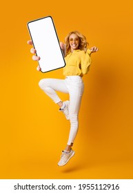 Cheerful Blonde Woman Jumping Up And Showing Newest Smartphone With Empty Screen, Enjoying New Application For Mobile Device. Orange Studio Background, Creative Collage With Mockup