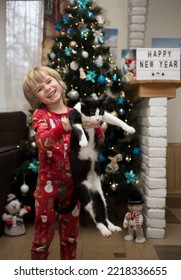 Cheerful Boy In Christmas Red Pajamas Holds A Domestic Black And White Cat In His Arms Stands At Home In Front Of The Christmas Tree. Concept Of Happy Cozy Family Winter Holidays Together With Pets