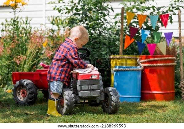 A cheerful boy\
dressed in a rustic style washes a tractor with a rag in the garden\
in the village. Playing in nature. The concept of raising a child\
and teaching them to work.