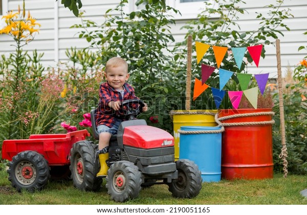 A cheerful boy dressed in a rustic style is\
sitting on a tractor in a garden in the village. Playing in nature.\
The child is interested in machinery and machines. The concept of a\
fun childhood.