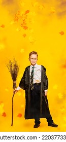 Cheerful Boy Dressed As A Wizard Stands With A Broomstick And Smiles. Full Length Portrait On A Yellow Background With Autumn Leaves. Happy Halloween! 