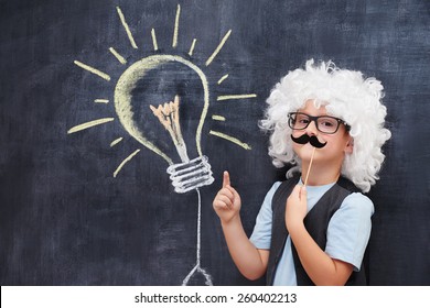 Cheerful Boy With Fake Mustache And Einstein  Wig Point On Bulb Visualized On The Blackboard