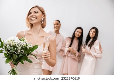 cheerful bride in wedding dress holding bridal bouquet and champagne glass, standing near blurred interracial bridesmaids on grey background, happiness, special occasion, blonde and brunette women - Powered by Shutterstock