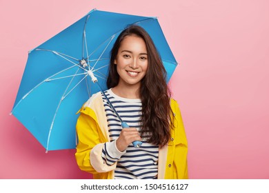 Cheerful brunette Asain woman with long dark hair, wears striped jumper, yellow raincoat, holds blue umbrella, has stroll during rainy day, isolated on pink. Bad weather doesnt spoil my mood - Powered by Shutterstock