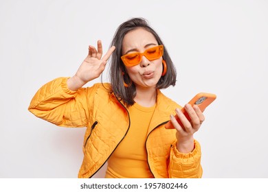 Cheerful brunette Asian woman dance to favorite song moves to rhythm of music holds modern smartphone wears trendy orange sunglasses stylish jacket expresses happiness and joy models indoor.
