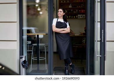 Cheerful brunette caucasian owner of coffee shop in apron standing at entrance greeting visitors and cliently hospitably, 20s smiling female barista in uniform opening cafeteria in morning - Powered by Shutterstock