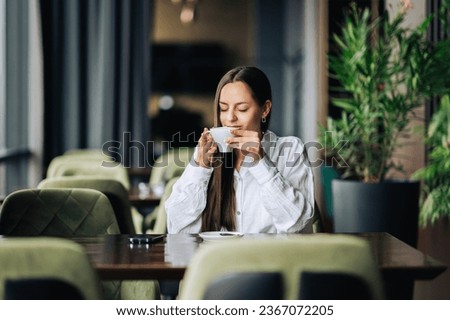 A cheerful brunette is sitting in a cafe and sipping fresh espresso and enjoying morning moments. A young trendy woman is enjoying her coffee in a cafeteria.
