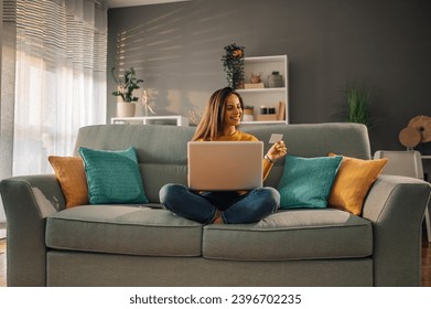 A cheerful brunette is sitting at her comfortable apartment with a laptop in her lap and credit card in the hand and purchasing online on the internet. A woman is using laptop for ordering online. - Powered by Shutterstock