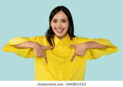 Cheerful brunette in vibrant yellow shirt playfully pointing downwards with both hands, positive young woman against trendy blue background. Promotions and attention-grabbing ads concept - Powered by Shutterstock