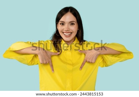 Cheerful brunette in vibrant yellow shirt playfully pointing downwards with both hands, positive young woman against trendy blue background. Promotions and attention-grabbing ads concept