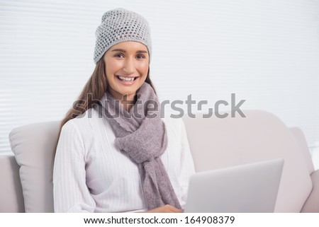 Cheerful brunette with winter hat on using her laptop sitting on cosy sofa