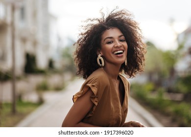 Cheerful brunette woman in brown dress smiles sincerely outdoors. Happy lady laughs and walks in great mood outside.