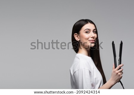 cheerful brunette woman holding hair straightener isolated on grey